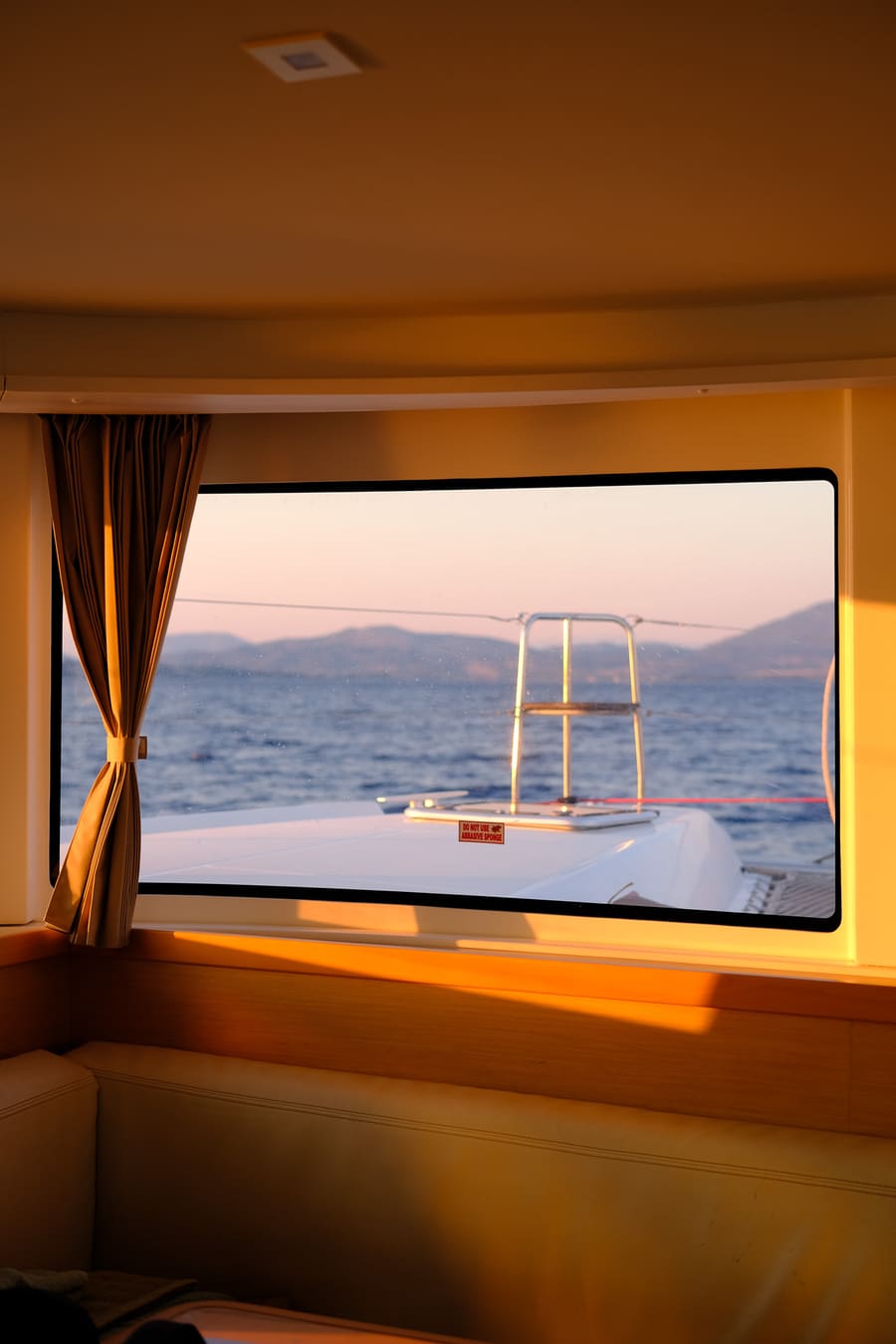 Golden-hour light through a yacht cabin window, looking out over the sea to distant hills